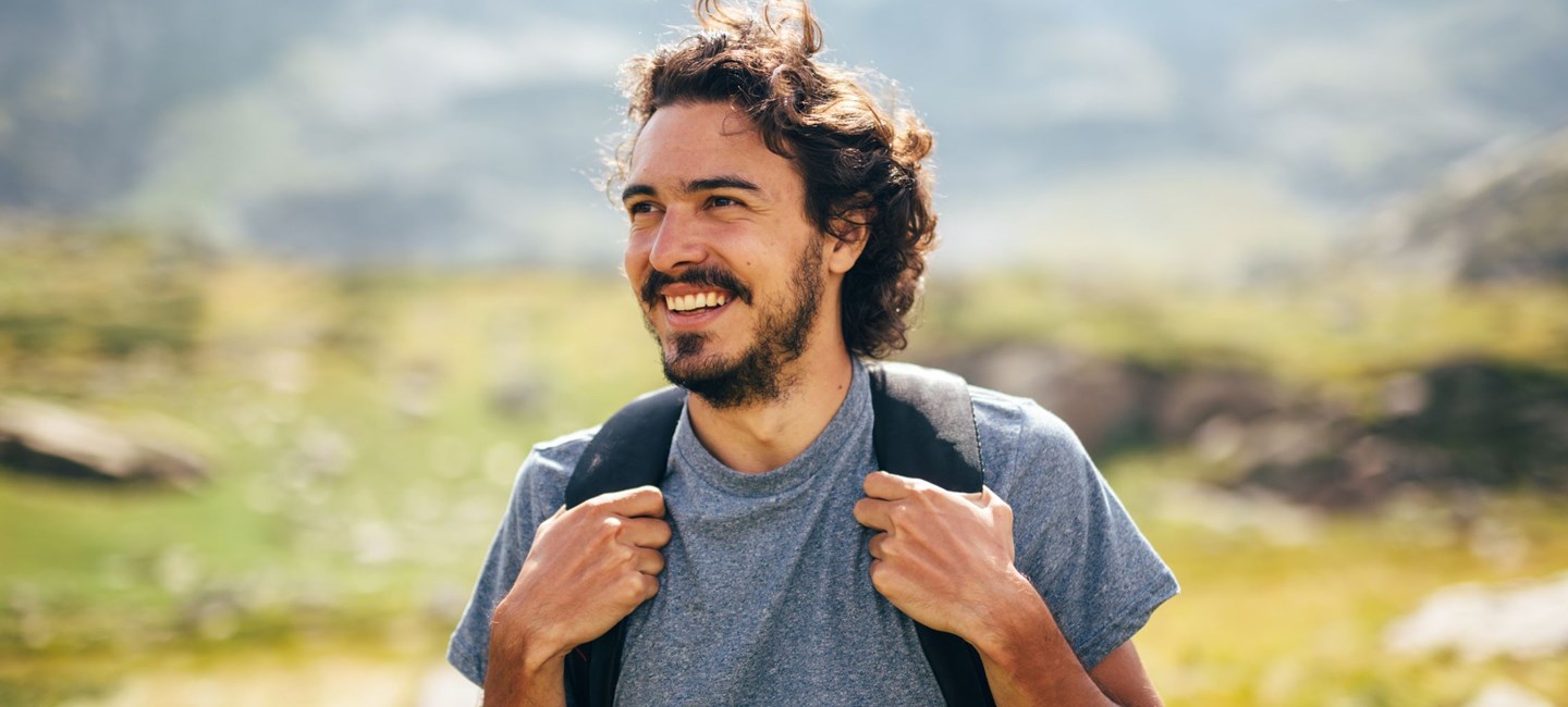 MAN IN A GREY T-SHORT WITH A BLACK BACKPACK ACROSS HIS SHOULDERS, STANDING OUTDOORS